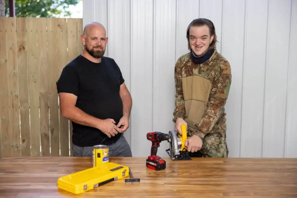 Two men standing together at a desk, one of them is holding a tool