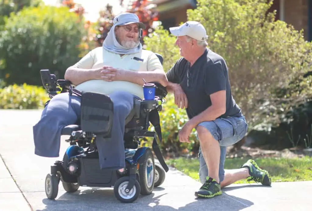 Two men chatting together, one is in a wheelchair, the other is kneeling down showing personal care