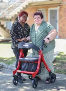 Two women standing together, one is holding a red zimmer frame