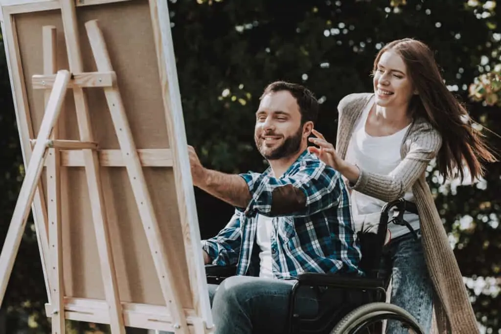 disabled young man in wheelchair drawing in a park with an women behind him, being supportive